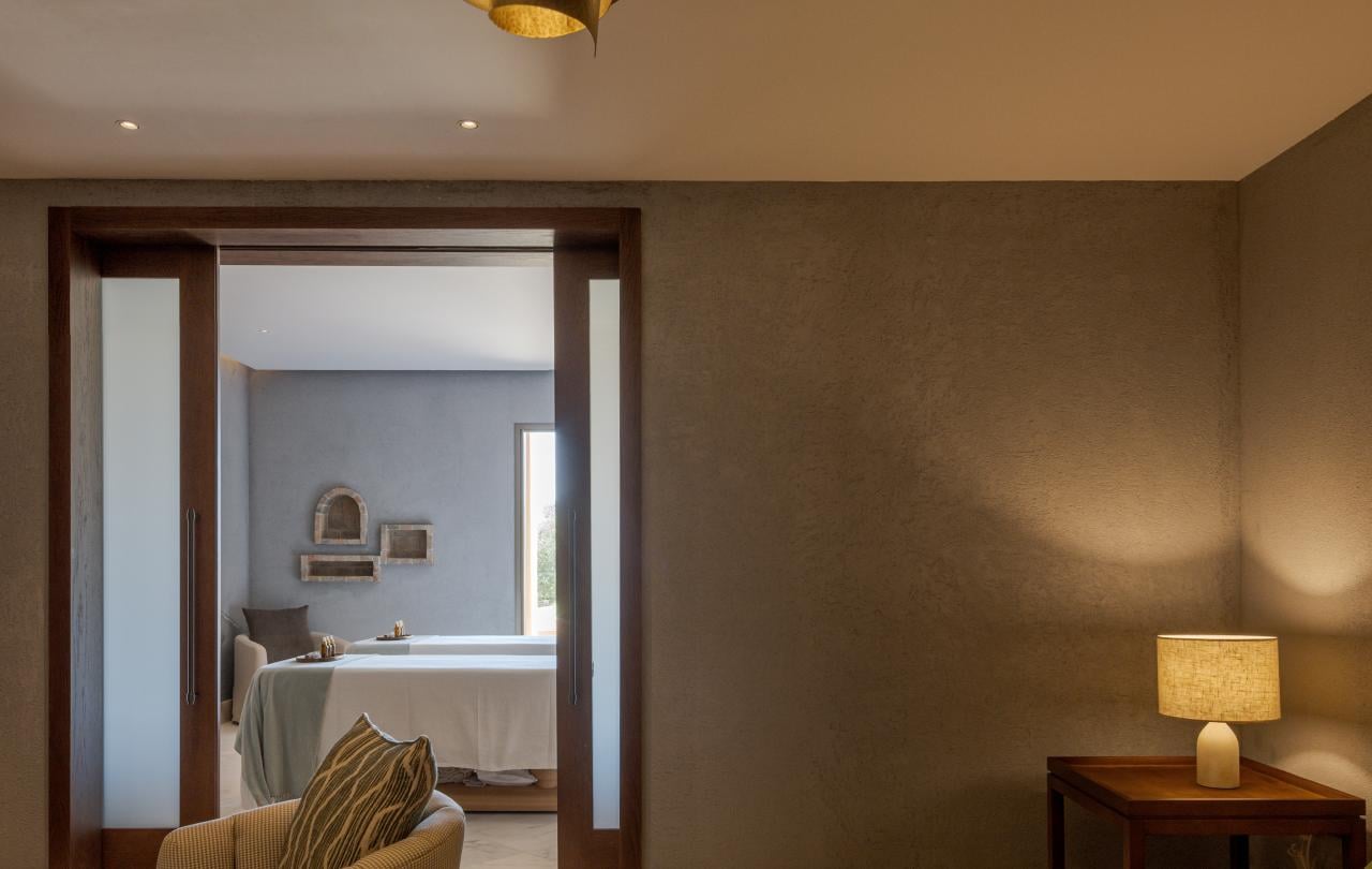 Sala de relaxamento acolhedora, com assentos confortáveis e um candeeiro de latão, com vista para uma sala de tratamentos.