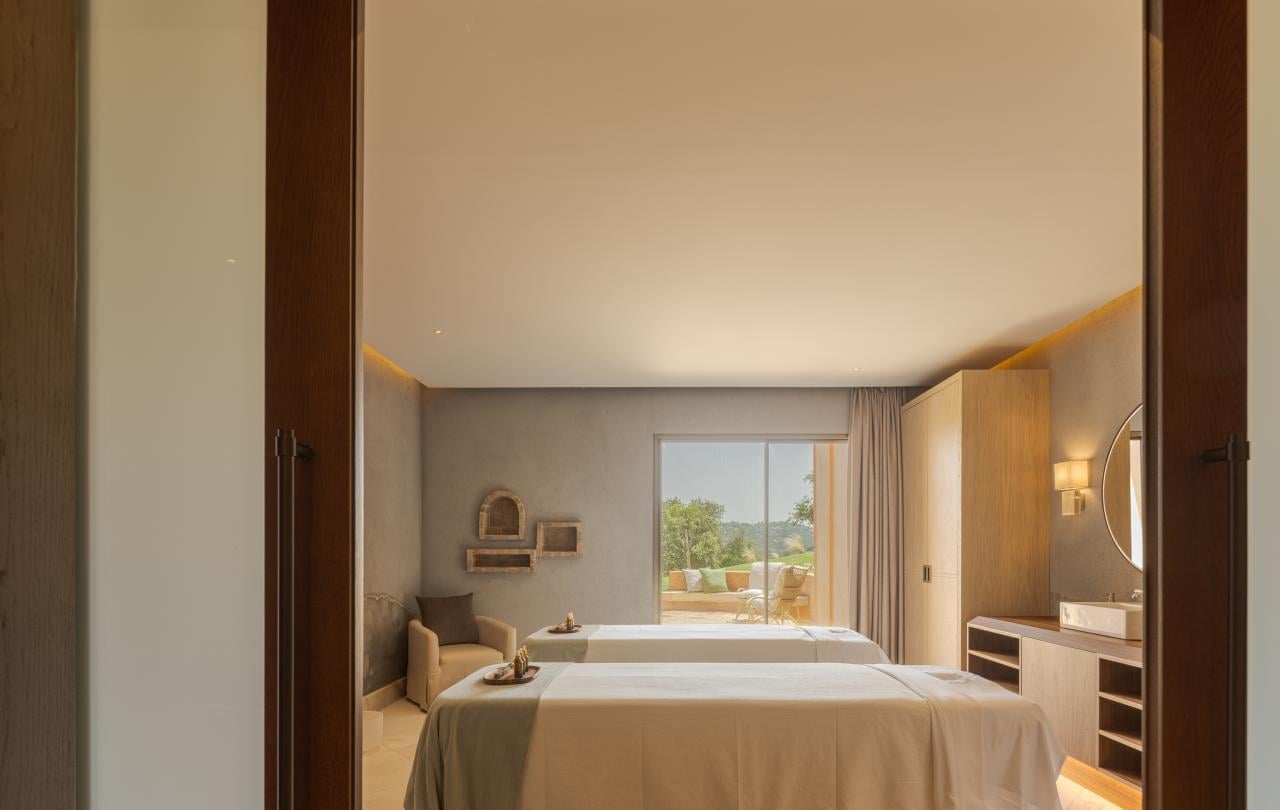 Vista emoldurada, através de portas de correr abertas, para uma moderna sala de tratamentos com duas camas de massagem e acesso ao terraço.