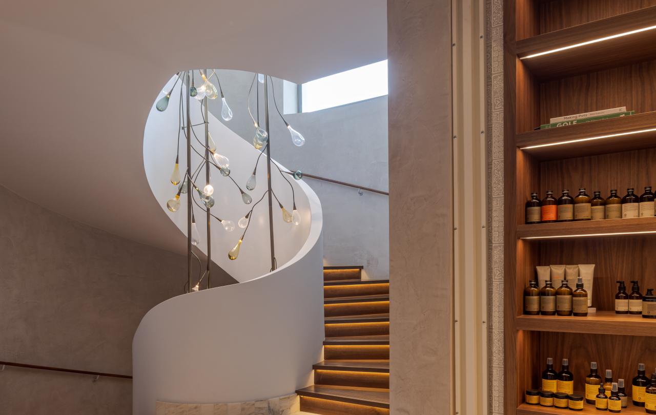 Imagem em grande plano de um lustre artístico em forma de lágrima suspenso no interior de uma estrutura em espiral que envolve uma escada branca.