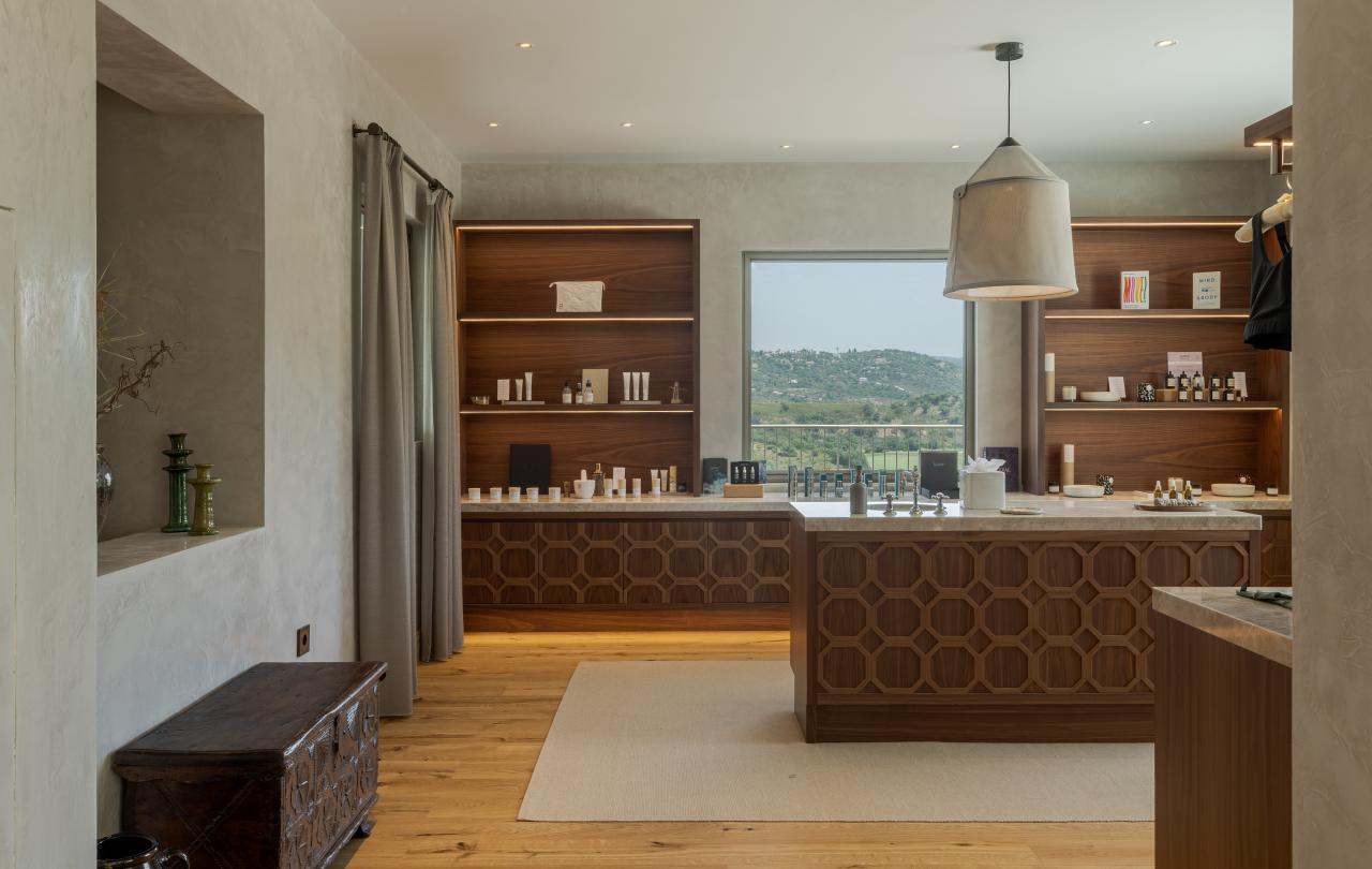 Boutique de spa com uma grande ilha central em madeira, prateleiras iluminadas e vistas panorâmicas pelo vidro para o exterior.
