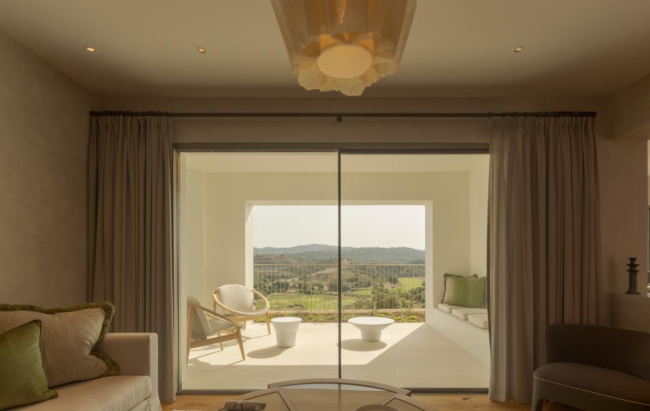 Elegante sala de estar interior com assentos confortáveis e uma mesa de centro redonda, com vista para um terraço ensolarado.