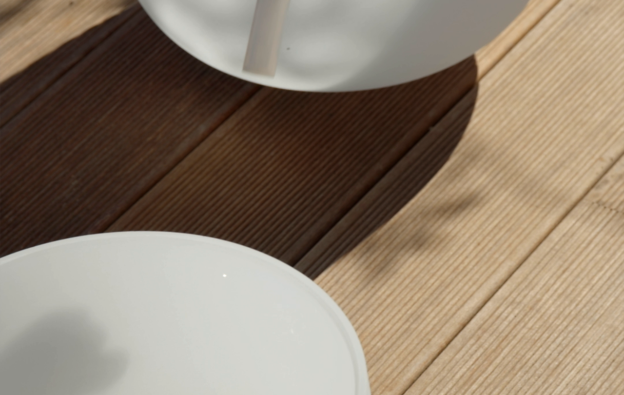 A hand holds a white mallet over a large white crystal singing bowl on a sunlit wooden deck, with a second bowl in the foreground.