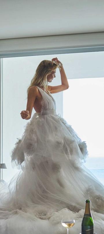 A bride poses in her wedding gown while standing on the bed of an oceanside hotel room