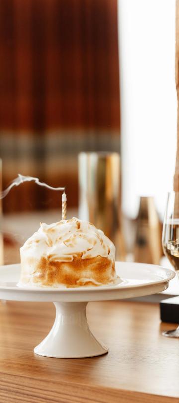 cake on white pedestal with candle recently blown out sits on table beside wine glasses