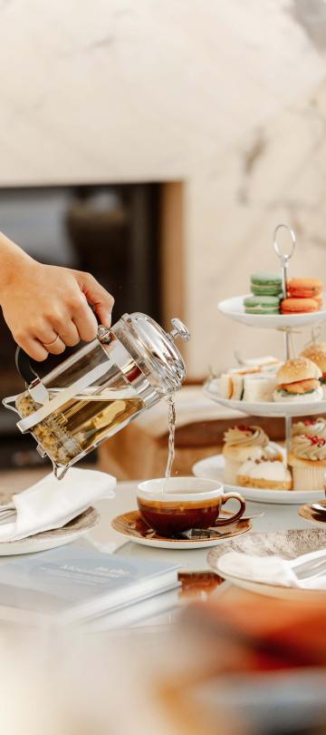 person pours tea from press into cup placed beside tiered dessert tray