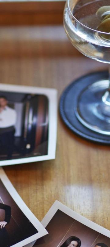 A scattering of three polaroid photos on a wooden desk top, with a camera, a half-full drink with an olive, a blurred privacy sign, and a small, black envelope with "Viceroy" embossed on it in gold.