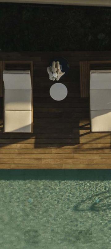 Seven lounging chairs with white padding lining the edge of a pool with high shadow. Small white tables sit between each pair of chairs.