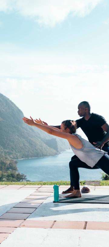 A man instructs a woman with a yoga pose on a terrace above a tropical beach at the foot of a mountain