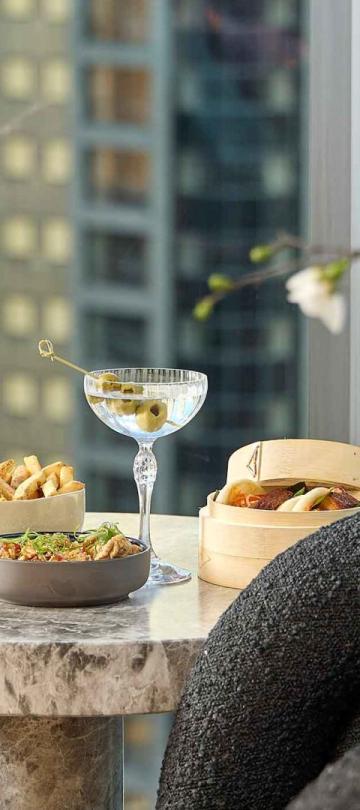 Two cocktails and snacks on a side table at Pandan, with a view of downtown Chicago through a window in the background