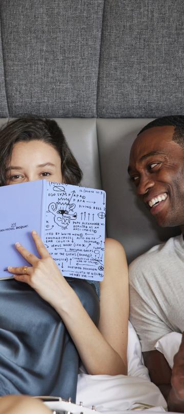 A woman holds a journal up to her face, hiding her smile as she lounges in a bed next to a smiling man holding a coffee cup.