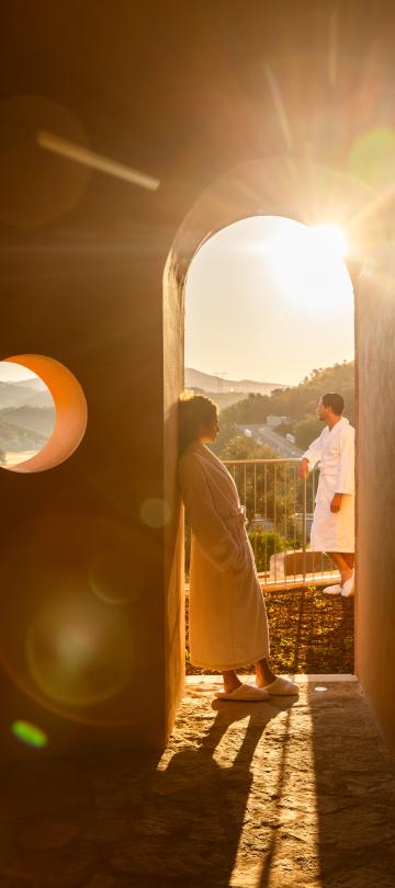 A couple in bathrobes looks out on the Algarve countryside as sunlight streaks through a rounded arch doorway