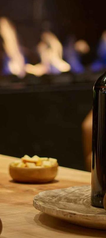 A bottle and glass of red wine on a table with a roaring fireplace in the background