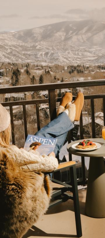A woman reads a magazine with her feet up, seated in a chair on a balcony overlooking a snowy Colorado mountain range