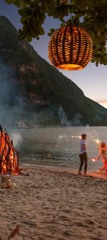 Three people dancing and playing with sparklers at a beach bonfire at night on Sugar Beach, in view of the Pitons