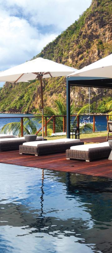 A row of pool recliners under patio umbrellas next to a pool, in view of the Pitons at Sugar Beach