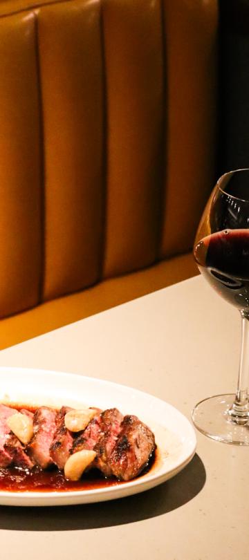 A steak dish accompanied by a glass of red wine, on a booth table at Dovetail