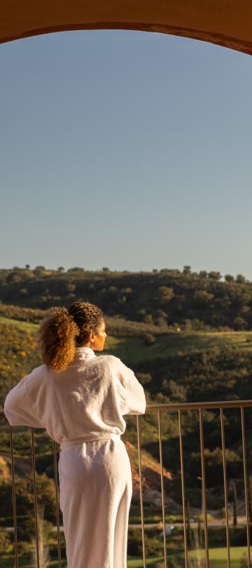 Woman in spa robe looks out over a balcony at rolling green hills.
