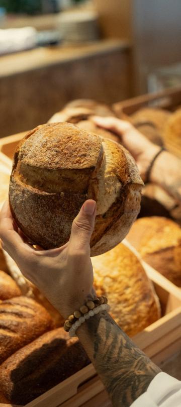 person holds loaf of bread over a shelf of different loaves of bread