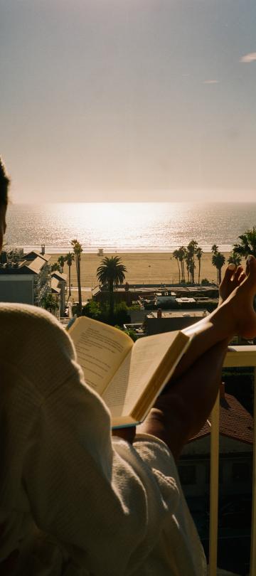 A person sits and reads with their feet up on the railing of their balcony, which overlooks the beach.