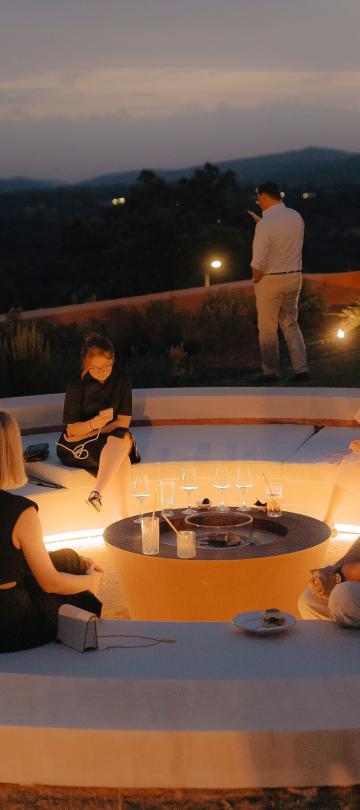 A group sits around a sunken fire pit at dusk.