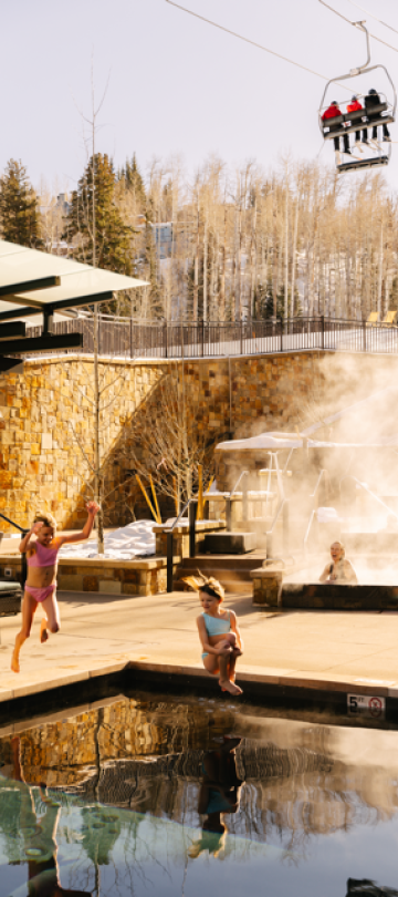 Two children play in a pool, while a ski lift with skiers passes by overhead.