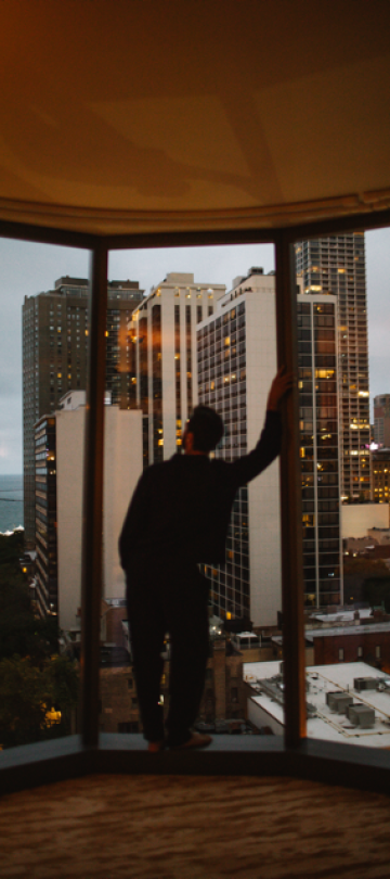 A man leans on the frame of the floor-to-ceiling windows of his hotel room, overlooking the city.