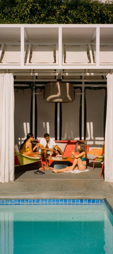 Three friends sit in a cabana, sipping drinks and talking.