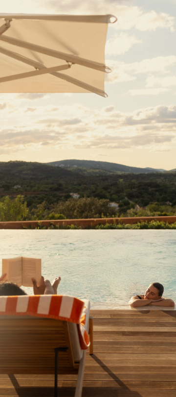 A person sits in a deck chair reading, while their partner lounges in the pool.