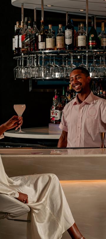 A bartender smiles at a patron seated at the bar, while shaking a cocktail