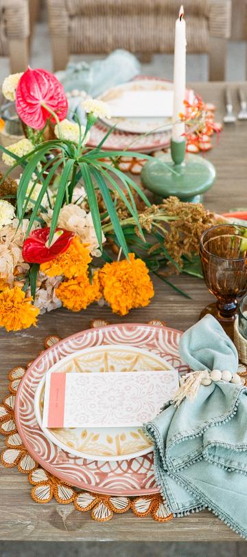 A colorful table setting features bright orange marigolds, red anthurium flowers, textured plates, a light blue napkin, and amber glassware on a rustic wooden table.