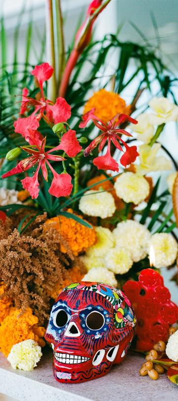 Brightly painted red sugar skull sits before a vibrant Day of the Dead altar arrangement featuring orange marigolds, red flowers, and green foliage next to a tall, molded brown candle.