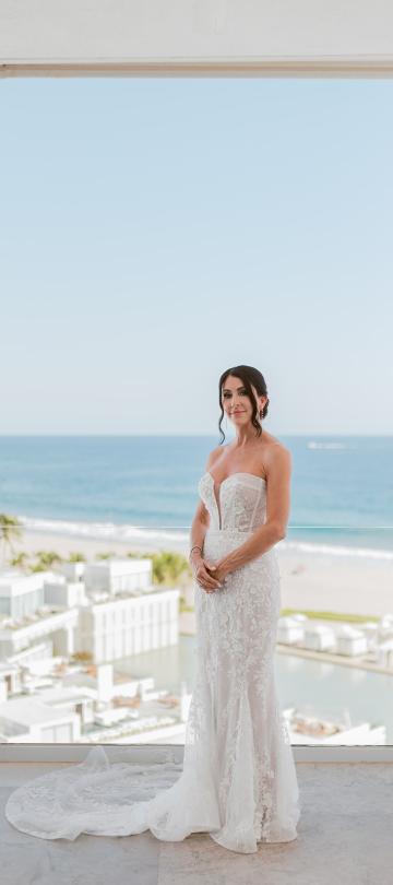 Bride in a white strapless lace wedding gown stands on a balcony overlooking a bright blue ocean, white resort buildings, and a beach below.