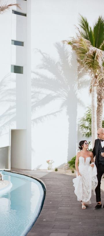A newly married couple in wedding attire walks along a dark pathway beside a bright blue pool, framed by a white building and tall palm trees casting distinct shadows.