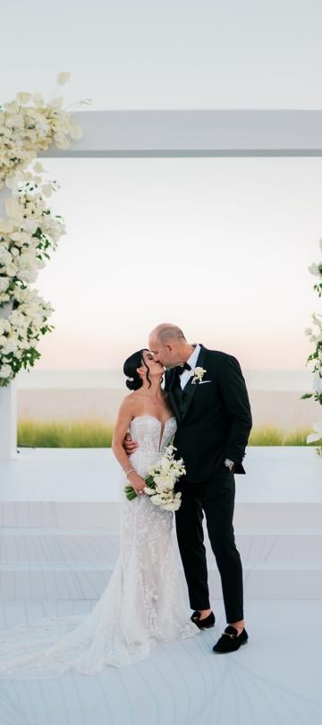 A newly married bride and groom kiss under a modern white arch decorated with cascading white floral arrangements during an outdoor ceremony overlooking the ocean.