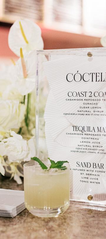 A clear acrylic cocktail menu titled "CÓCTELES" stands next to a glass of pale green iced drink garnished with mint, with white floral arrangements blurred in the background.