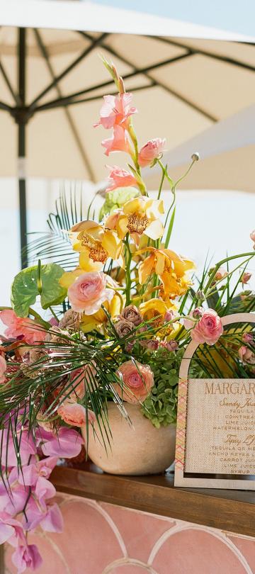 Lush floral arrangement featuring yellow orchids, pink roses, and tropical greenery next to a wooden-framed margarita menu sign on a pink tiled outdoor bar under a large umbrella.