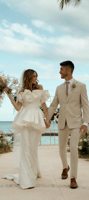 Bride and groom walking hand in hand on a beachside terrace, surrounded by palm trees and elegant floral arrangements. 