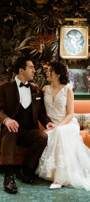 A newly married couple in formal attire sits close together on an orange sofa against a dark, richly patterned botanical wallpaper in a moodily lit room.