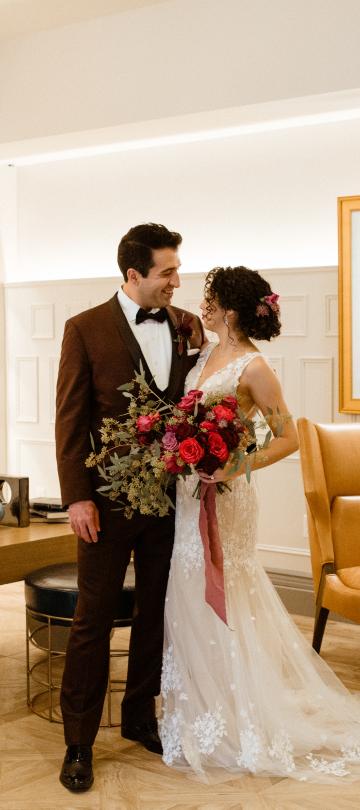 A newly married couple stands close together in a stylish, brightly lit room featuring built-in bookshelves, modern art, and tan leather seating.