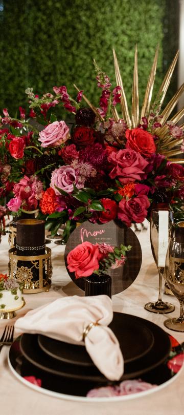 A richly decorated table setting features a dramatic centerpiece of deep red and pink roses against a green hedge backdrop, complemented by gold accents and dark tableware.