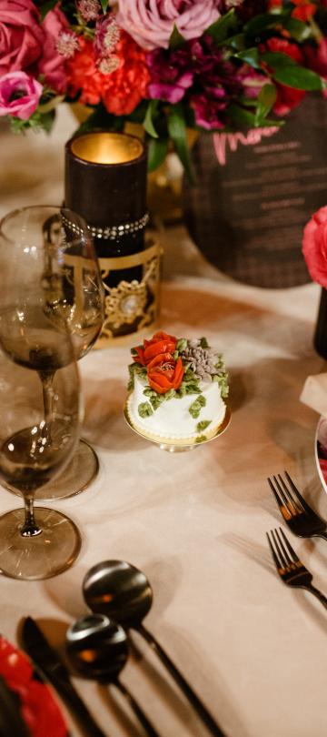 A formally set table features tinted wine glasses, black cutlery, a small white cake with red and green floral frosting, a lit dark candle, and a vibrant pink and red floral centerpiece.