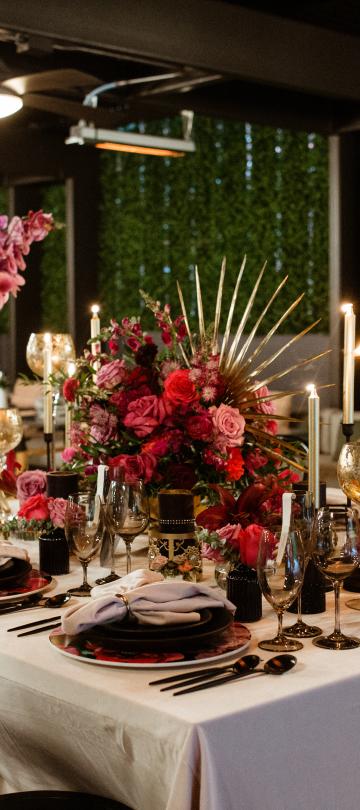 Uma elegante disposição de mesa para um jantar formal, com centros de mesa florais em vermelho intenso, luz de velas e louça preta, tendo como pano de fundo um pátio escuro com sebes verdes.