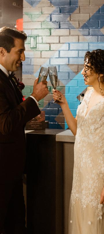 A smiling groom in a brown suit and a bride in a white lace gown toast with champagne flutes in front of a colorful geometric brick wall.