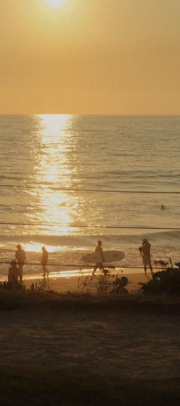 Las siluetas de unos bañistas y un surfista con su tabla se recortan en la orilla al atardecer, mientras el sol se refleja con intensidad en las aguas del océano.