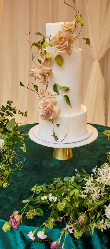 Three-tiered white wedding cake adorned with blush sugar roses and trailing vines, set on a gold stand atop a dark emerald velvet draped table surrounded by lush green and white floral arrangements.