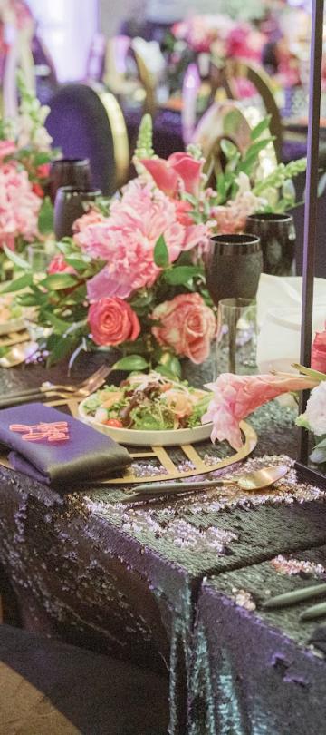 Uma mesa de banquete ricamente decorada apresenta uma toalha preta com lantejoulas, centros de mesa altos com arranjos florais em tons de rosa e vermelho, talheres dourados e guardanapos escuros com bordados coloridos.