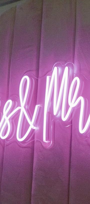 Um letreiro em neon rosa vivo com a inscrição «Mrs & Mrs» ilumina um pano de fundo drapeado, com textura e em tons de rosa-malva.