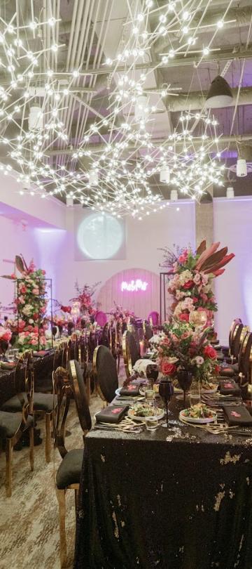 Um espaço para eventos luxuoso, com mesas decoradas com lantejoulas pretas, arranjos florais altos com flores cor-de-rosa e vermelhas, cadeiras com detalhes dourados e uma instalação suspensa espetacular composta por luzes brancas interligadas.