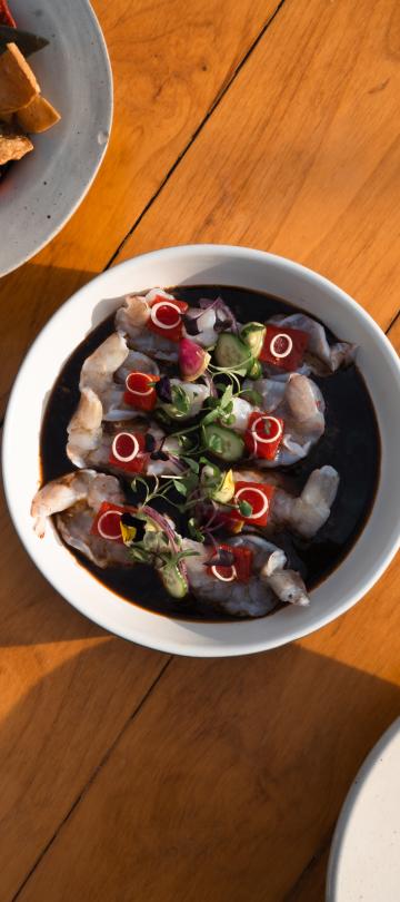 Overhead view of shrimp ceviche in dark sauce with vegetable garnishes, served on a wooden table next to a plate of tortilla chips.
