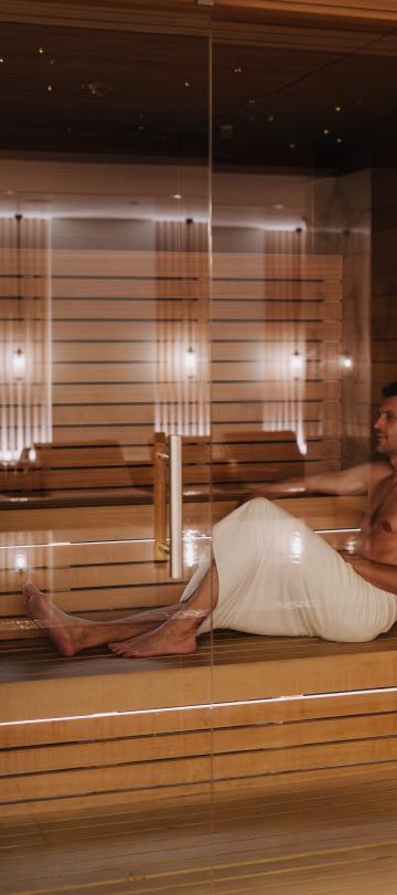 Man in a white towel relaxes on a wooden bench in a modern sauna.